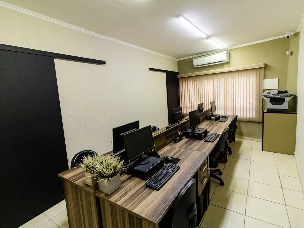 Sala de trabalho bem iluminada com mesas de madeira enfileiradas, computadores, cadeiras de escritório e pequenos vasos de plantas, demonstrando a infraestrutura operacional do escritório de contabilidade.