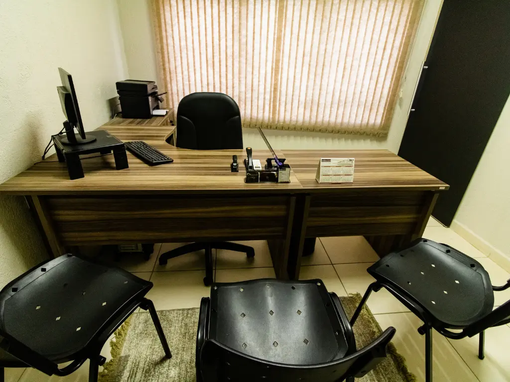 Sala de trabalho bem iluminada com mesas de madeira enfileiradas, computadores, cadeiras de escritório e pequenos vasos de plantas, demonstrando a infraestrutura operacional do escritório de contabilidade.